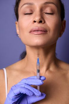 Close-up beauty portrait of middle aged beautiful woman posing on purple background with a syringe of cosmetic rejuvenating product in her hand. Injection cosmetology, anti-aging treatment concept
