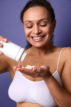 Smiling cheerful African woman in white underwear holds a bottle of food supplement and pours a vitamin complex into her hand. Medical pills for healthy skin. Health care and medicine concept