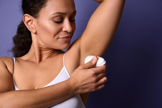 Close-up of a beautiful woman applying roll-on deodorant underarm. Protective sweating cosmetic product, personal hygiene, grooming self-care concept on violet background with copy ad space