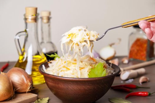 Fermented cabbage in ceramic bowl on table with spices and ingredients. Hand holds fork with salad. Healthy food, Russian traditional cuisine. Selective focus.