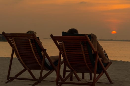 A close-up of two tourists on sunbeds in front of a beautiful sunset on a tropical island. Impressive image for any use.