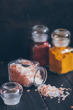 Bright aromatic spices in glass jars, assortments of spices: pink salt, paprika and turmeric on black stone background