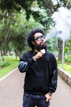 Vape. Young brutal man with large beard and fashionable haircut in sunglasses smoking an electronic cigarette in the city park. Steam cloud. Lifestyle.