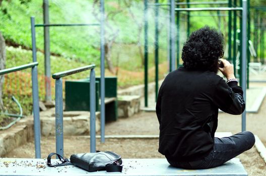 Man with a beard smokes an electronic cigarette sit on a bench in the park