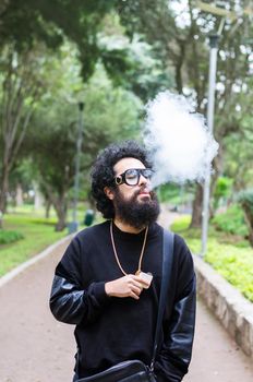 Vape. Young brutal man with large beard and fashionable haircut in sunglasses smoking an electronic cigarette in the city park. Steam cloud. Lifestyle.