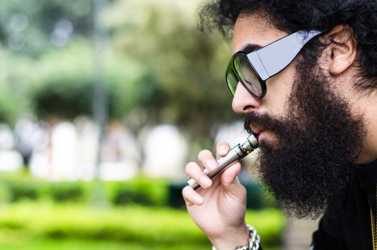 Bearded man smokes vape close up. Electronic cigarette concept. Man with long beard and clouds of smoke looks relaxed. Man with beard and mustache on calm face, branches on background, defocused.