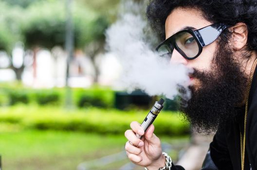 Bearded man smokes vape close up. Electronic cigarette concept. Man with long beard and clouds of smoke looks relaxed. Man with beard and mustache on calm face, branches on background, defocused.