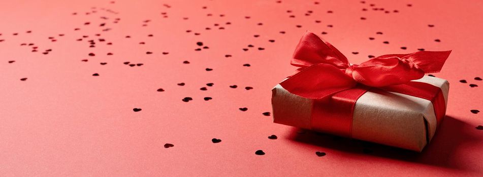 Valentines Day. Craft Gift box with beautiful red ribbon with heart shape confetti on red background with copy space. Banner