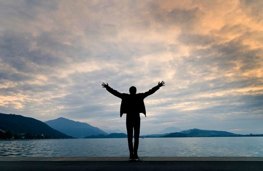 Silhouette of a man with his back to camera, arms outstretched in front of a landscape of a lake at sunset with a sky with clouds colored by sunlight.