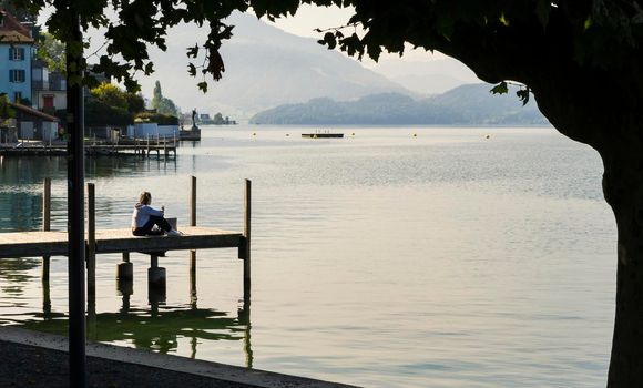 View of calm Lake Zugersee in the Swiss town of Zug with a small wooden harbor and a young girl sitting looking out over the lake at sunrise.