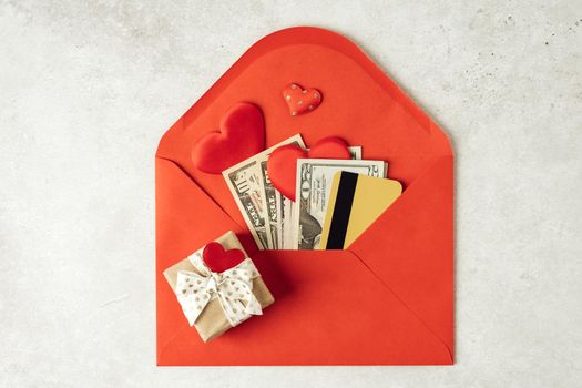 Red paper envelope with money dollars bills and credit card. Flat lay of gray working table background with Valentine gift, letter, heart shape. Top view, mock up. Valentine's Day budget