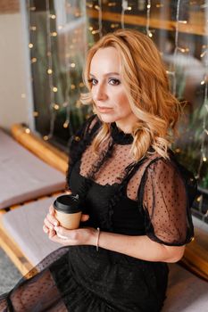 Young charming blonde with a cute smile and makeup while relaxing in a cafe. She is holding a cup of coffee in her hands. She is dressed in a black dress with transparent sleeves