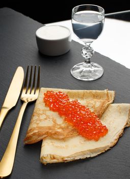 Golden russian pancakes with red caviar and sour cream and a glass of vodka isolated on black background. Russian national cuisine.