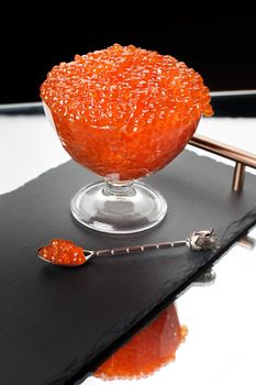 Red caviar in a glass bowl with a silver spoon on a tray. Black background, mirror reflection