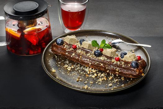 Eclair with chocolate, berries and halva served with tea on dark background. Tasty dessert. Sweet Dessert Pastry filled with cream. Chocolate icing.