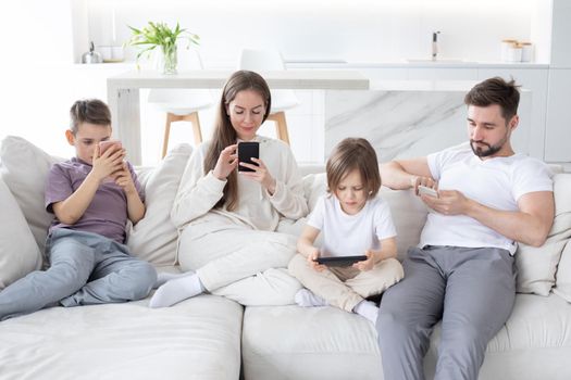 Family of parents and children sitting together at sofa with smartphones at home