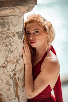 Outdoor portrait of a young beautiful natural redhead girl with freckles, long curly hair, in a red dress, posing against the background of the sea