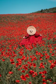 Young woman stands with her back in a long red dress and hat, posing on a large field of red poppies.