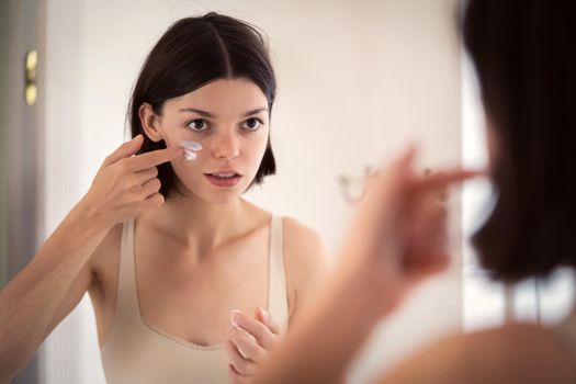 A young pretty girl applies cream stripes on the cheeks, declaring war on dry skin and acne. A woman takes care of the health and hydration of her skin in the bathroom near the mirror.