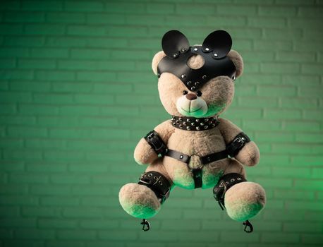 the toy Teddy bear dressed in leather belts and mask accessory for BDSM games on a light background texture of a brick wall