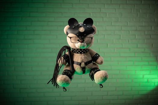 the toy Teddy bear dressed in leather belts and mask accessory for BDSM games on a light background texture of a brick wall