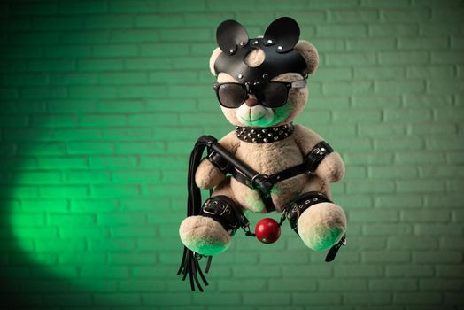 the toy Teddy bear dressed in leather belts and mask accessory for BDSM games on a light background texture of a brick wall