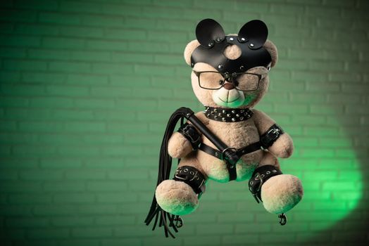 the toy Teddy bear dressed in leather belts and mask accessory for BDSM games on a light background texture of a brick wall