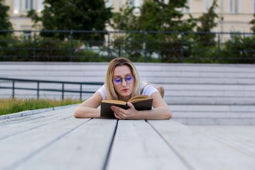 Young woman hipster reading a book lying on the bench in summer park, horizontal