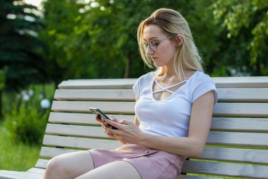 Young woman in eyeglasses sitting on the bench uses a smartphone in summer park, horizontal