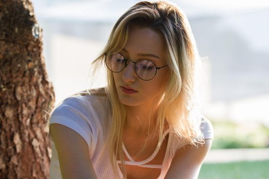 Portrait of young attractive woman in eyeglasses near the tree in park at sunny day, horizontal