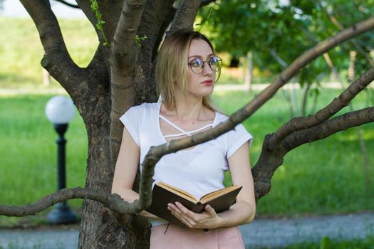 Attractive young woman with a book in the hands in the park leaning on a tree branch, horizontal