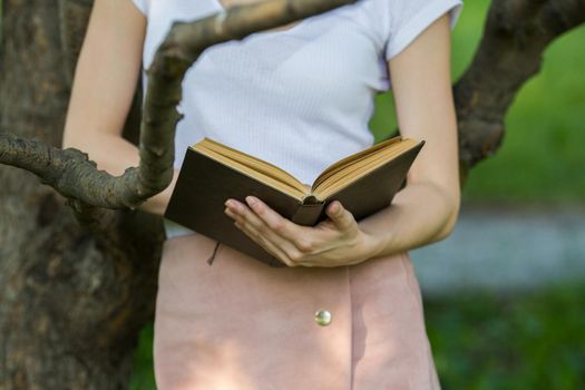 Young woman holding a book in hands leaning on a tree branch, horizontal