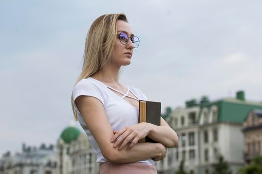 Young melancholic woman with a book in her hands standing at cityscape on background, horizontal