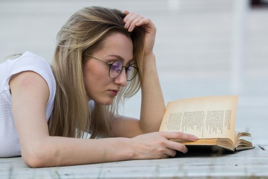 Portrait of young woman reading a book lying on the bench in the park, close up