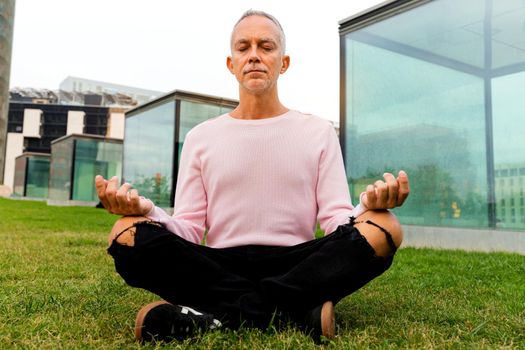 Caucasian adult man wearing pink sweater meditating in city public park. Meditation and spirituality concept.
