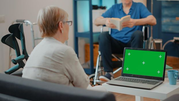 Retired person with crutches looking at laptop with green screen in living room. Old woman with disability looking at computer with isolated mock up template and blank chroma key background.