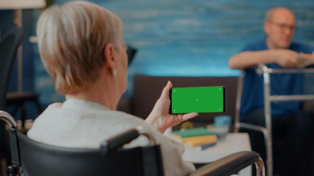 Retiree with disability using horizontal green screen on mobile phone. Elder woman in wheelchair looking at blank copy space with isolated chroma key and mock up template on smartphone.