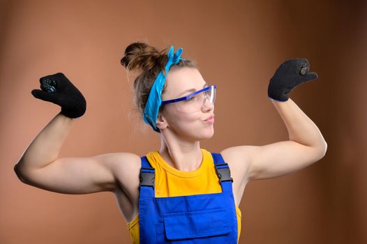 Strong stylish caucasian woman repairman foreman in overalls showing a gesture of strength showing biceps. Studio portrait of a young and strong feminine woman.