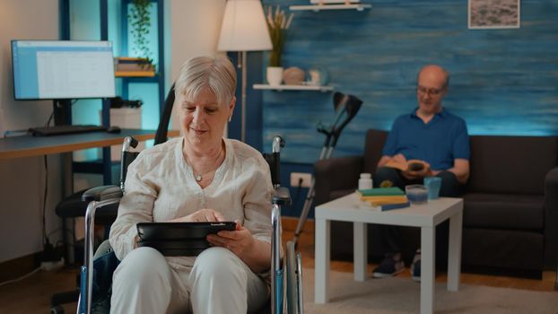 Retired person using digital tablet and sitting in wheelchair. Elderly woman with chronic disability looking at device to browse internet. Grandma using mobility support and gadget.