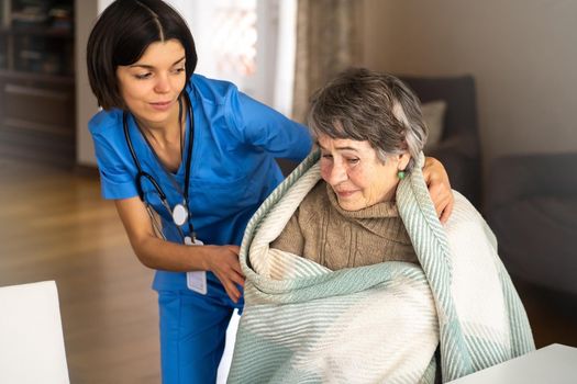 A young nurse takes care of an elderly 80-year-old woman at home, wraps a blanket around her. Happy retired woman and trust between doctor and patient. Medicine and healthcare.