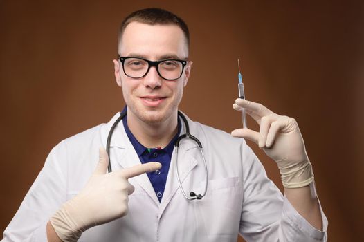 A young Caucasian doctor in a white coat and glasses holds a syringe with a vaccine. Friendly cuddly young man points a finger at a syringe offering to be vaccinated.