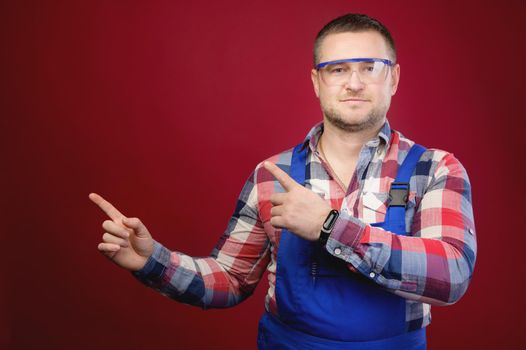 Attractive Caucasian male foreman in uniform and goggles points fingers at blank space. Foreman repairman maste studio portrait. on red background