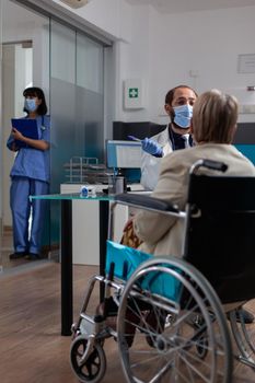 Physician meeting with old patient in wheelchair at checkup during covid 19 pandemic. Specialist giving support and advice to pensioner with physical disability in medical office.