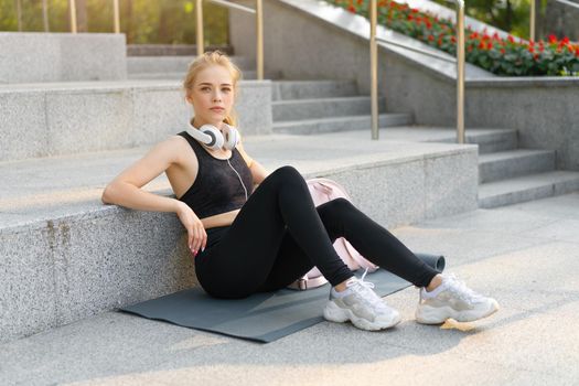 Sport and Fitness Young Adult Caucasian Woman Sitting Exercise Mat Concrete Floor Resting after morning Workout Outdoor Summer Park Athletic Female Take a Break listen Music Headphones