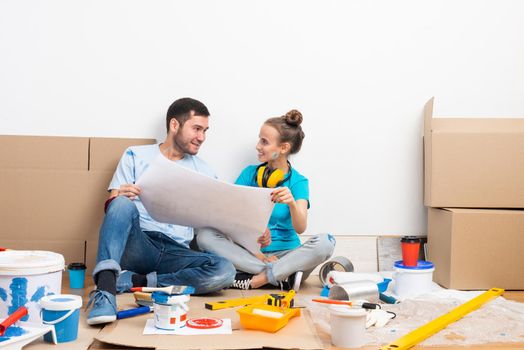 Happy couple sitting on floor with construction blueprint. Home remodeling and house interior redesign. Construction tools and materials lying on floor. Young family studies renovation project of flat