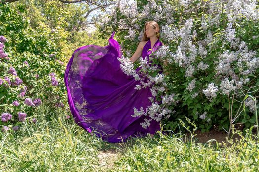 Fashion Model in Lilac Flowers, Young Woman in Beautiful Long Dress Waving on Wind, Outdoor Beauty Portrait in Blooming Garden.