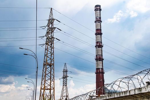 High voltage tower with wires and industrial chimney. High-voltage transmission line of electrical energy. Chimney of the enterprise. Industrial landscape. Cloudy sky horizon. Background.