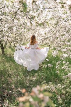 Fantasy woman in long white elegant fashion long dress walks in green spring blossom cherry garden. Happy cheerful girl princess bride. Skirt fabric flies flowing waving in wind motion