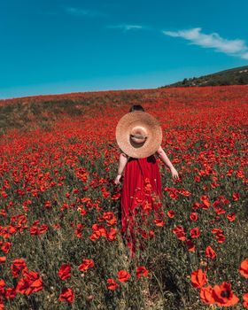 Young woman stands with her back in a long red dress and hat, posing on a large field of red poppies.