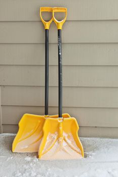 Two yellow and black snow shovels are dusted in snow and leaning against a wall ready for use. One shovel is wider than the other one.